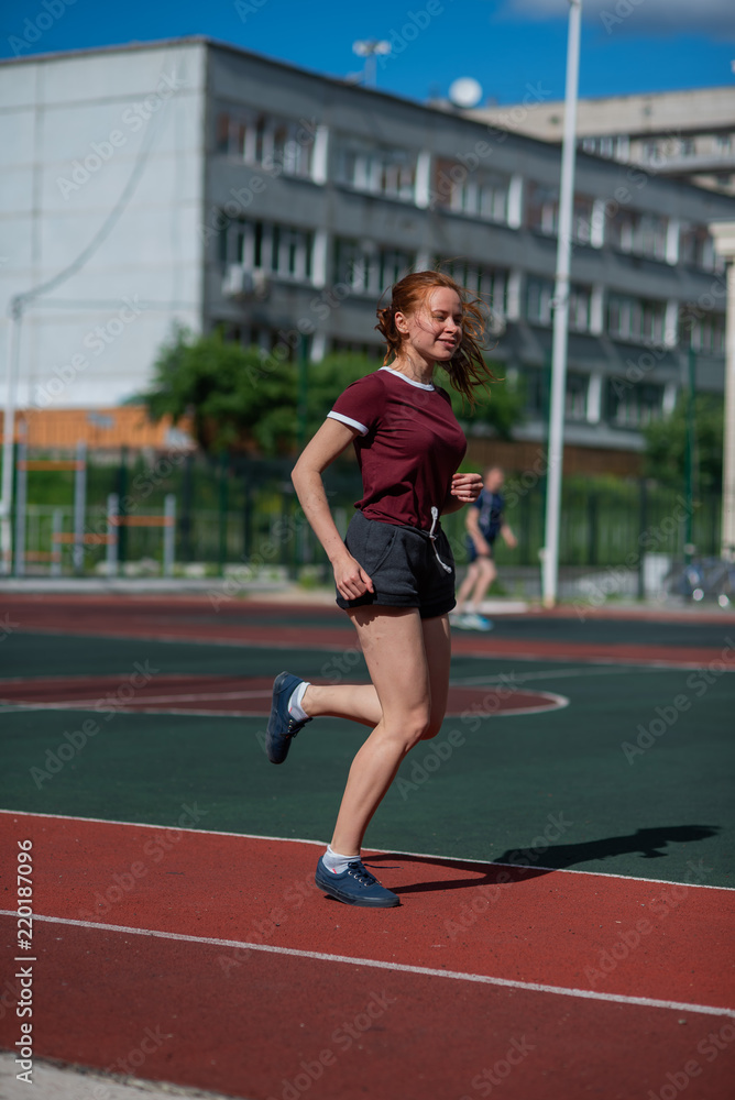 Red-haired young girl runs in the stadium. Student delivers standards for running outdoors. Young woman running on the football field