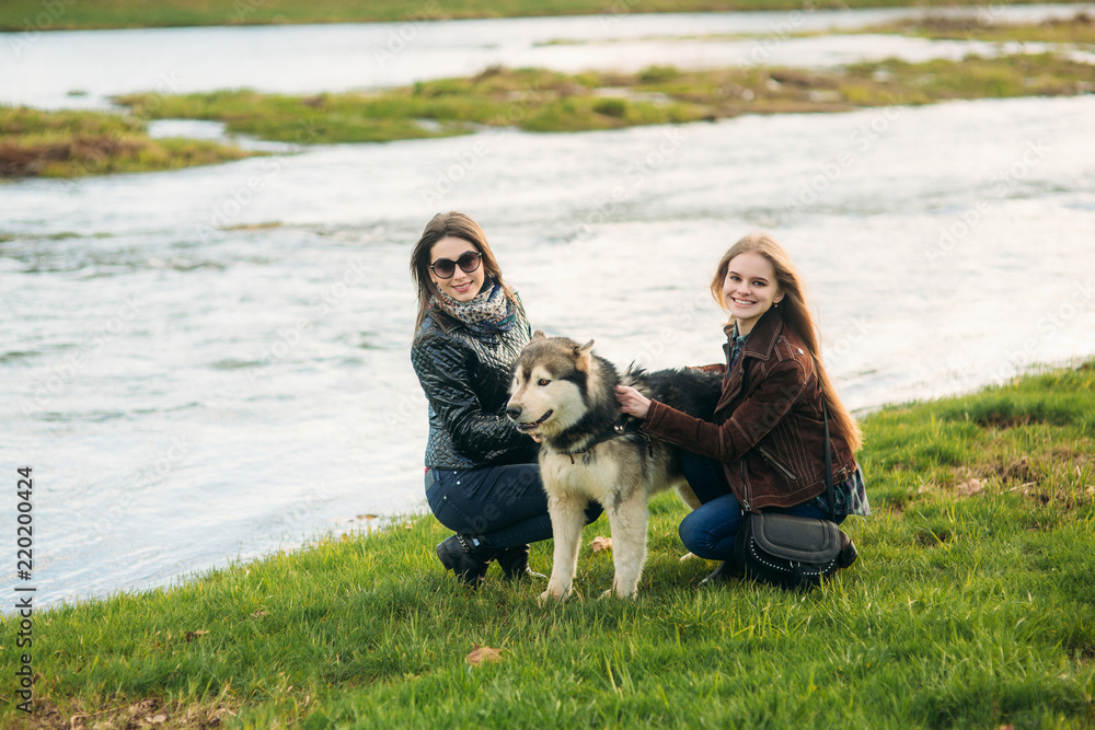Two sisters walk with husky dog in front of river. Brunettle and blonde girl and caress a dog