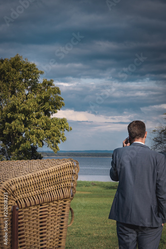 Businessman or manager, a young man is standing in a park by the lake talking on the phone with a smartphone. He is wearing a suit, his eyes are on the lake. He is standing next to a beach chair