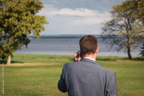 Businessman or manager, a young man is standing in a park by the lake talking on the phone with a smartphone. He is wearing a suit, his eyes are on the lake