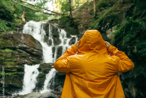 man standing in yellow raincoat and looking at waterfall