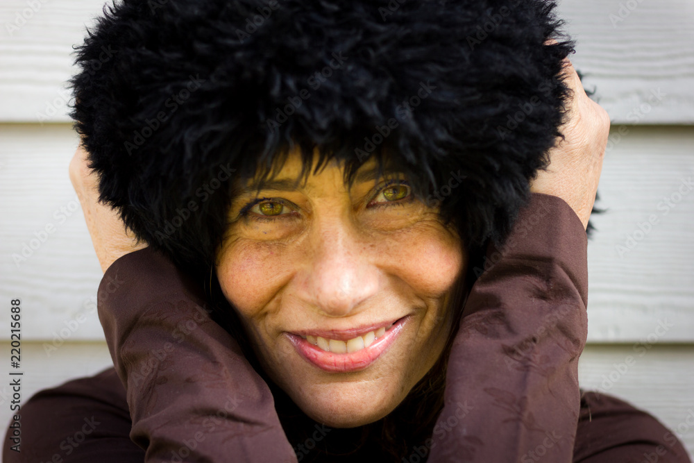 Pretty mature lady ready for cold weather. Close up of gorgeous middle aged woman with green eyes and hands on black fur Russian winter hat on head over white wood plank background. Winter fashion Pretty mature lady ready for cold weather. Close up of gorgeous middle aged woman with green eyes and hands on black fur Russian winter hat on head over white wood plank background. Winter fashion