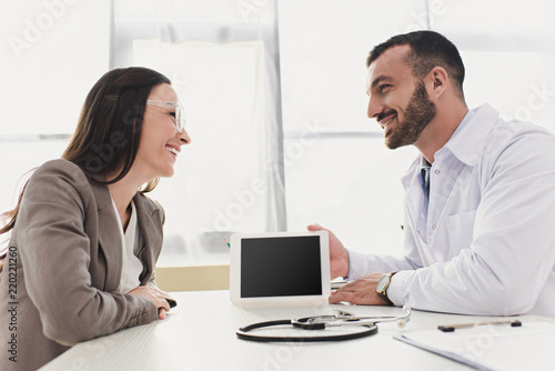smiling doctor showing patient tablet with blank screen in clinic