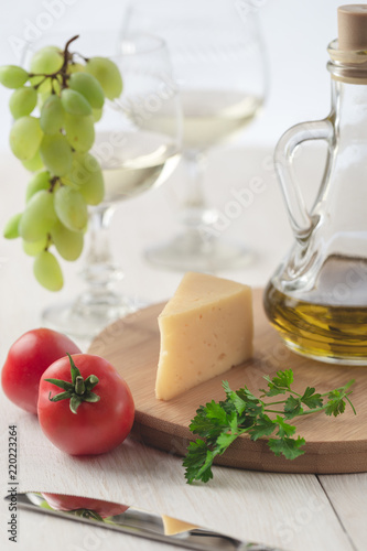 On the table is cheese, tomatoes, grapes, olive oil, parsley, two wineglasses and a bottle of white wine. Also on the white table is a cutting board and a knife.