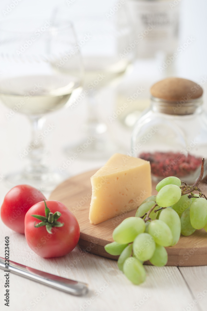 On the table is cheese, tomates, grapes, olive oil, parsley, two wineglasses and a bottle of wine.