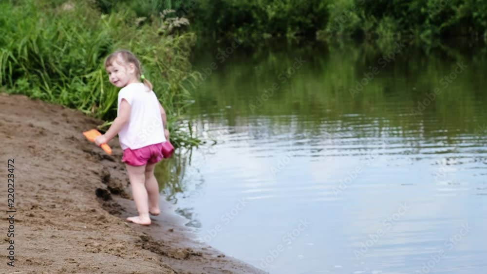 Little girl is walking with a toy spatula and a bucket in the sand near the river Little girl is walking with a toy spatula and a bucket in the sand near the river