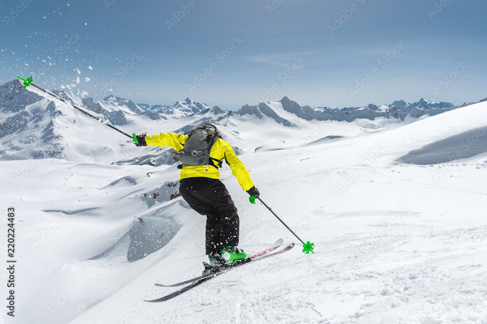 A skier in full sports equipment jumps into the abyss from the top of the glacier against the background of the blue sky and the Caucasian snow-capped mountains. View from the back. Elbrus region