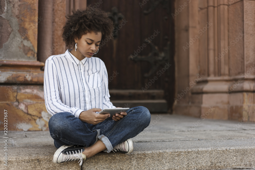 African-American girl sitting on the steps near the building. She works on a tablet. Using tablet. African-American girl sitting on the steps near the building. She works on a tablet. Using tablet.