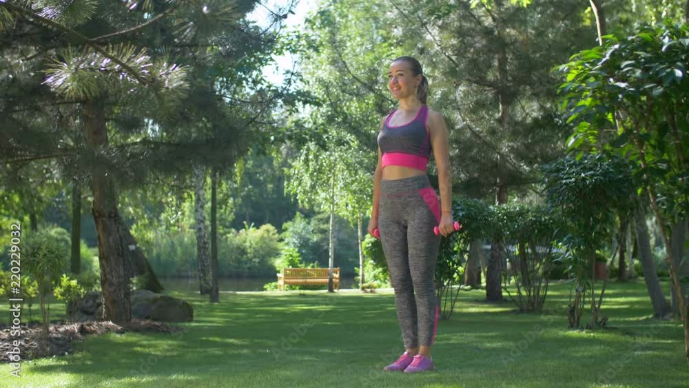 Attractive sporty fitness woman with ponytail doing squats and holding dumbbells while standing on green grass in summer park. Positive fit female with dumbbells squatting during workout outdoors. Attractive sporty fitness woman with ponytail doing squats and holding dumbbells while standing on green grass in summer park. Positive fit female with dumbbells squatting during workout outdoors.