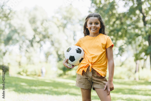 beautiful happy kid holding soccer ball and smiling at camera in park