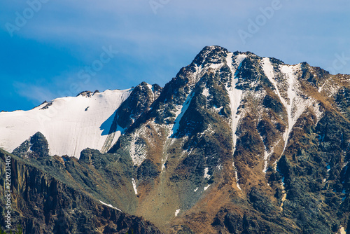 Snowy mountain top in blue clear sky. Rocky ridge under clouds in sunny day. Atmospheric minimalistic landscape of majestic nature.