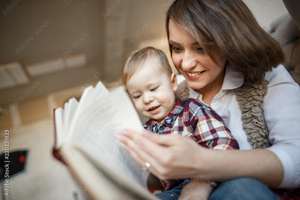 Mother is sitting with her baby and reading a book. She is smiling and looking at it. Baby is sitting on mom's lap and looking at book as well. Mother is sitting with her baby and reading a book. She is smiling and looking at it. Baby is sitting on mom's lap and looking at book as well.