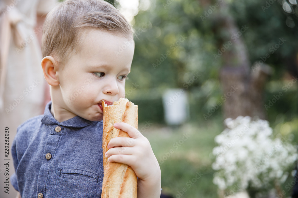 A picture of boy standing outside and holding baguette. He is biting a piece from it. Kid is looking backwards. A picture of boy standing outside and holding baguette. He is biting a piece from it. Kid is looking backwards.