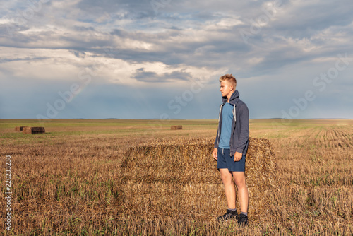 a wandering guy in a hoodie in a hood looks thoughtfully at a wheat field with a backdrop of the setting sun of a stormy sky © Tatsiana