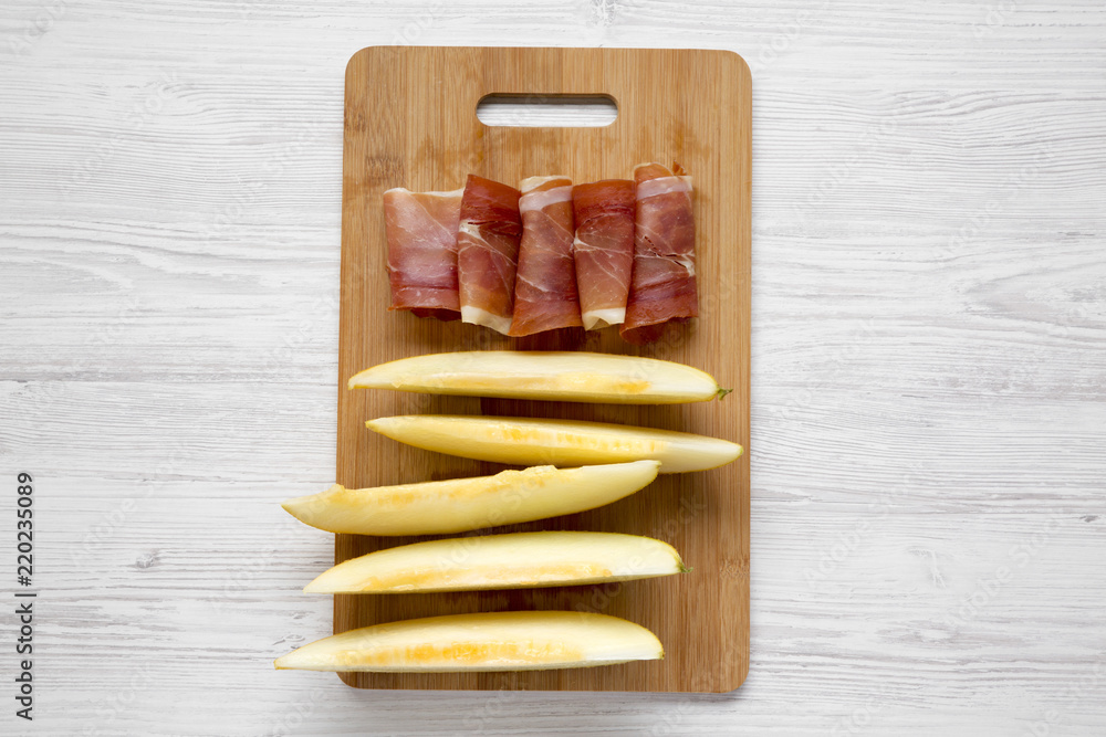 Melon slices with prosciutto on bamboo board over white wooden surface, top view. Overhead, flat lay, from above.