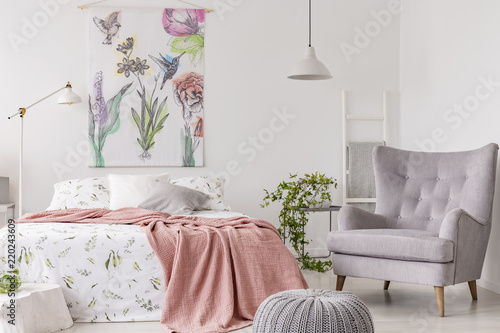 A sunny bedroom interior with a bed dressed in green pattern white linen and a peach blanket. Gray comfortable armchair beside the bed and a textile print of flowers and birds above. Real photo.