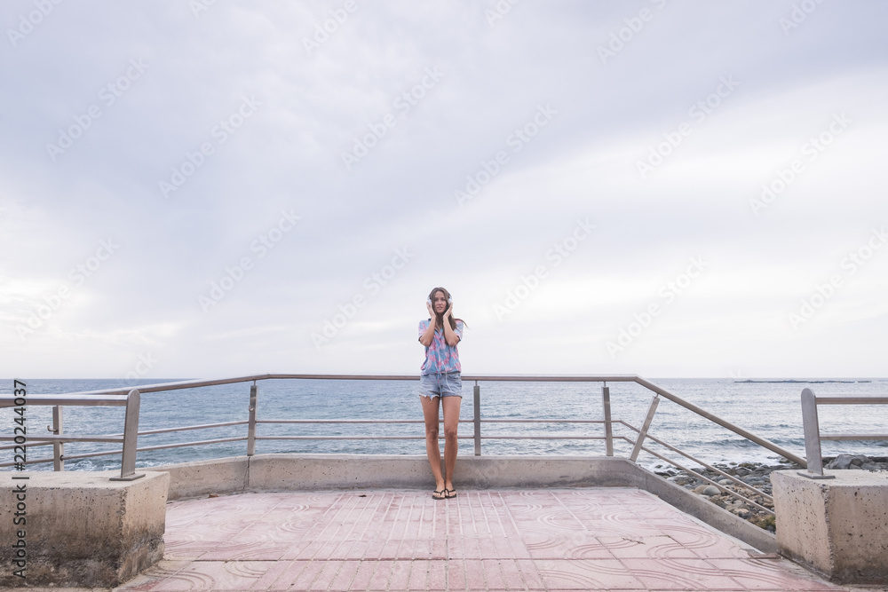 Full length portrait of beautiful young woman standing with the ocean and water blue in background and sky listening music with headphones. modern concept and leisure activity for nice girl