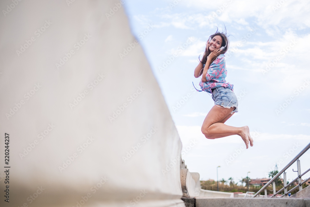 Happiness concept with young beautiful girl jumping with joyful against a white sky blue background. sea side place. Cheerful caucasian woman smile and have fun listening music Happiness concept with young beautiful girl jumping with joyful against a white sky blue background. sea side place. Cheerful caucasian woman smile and have fun listening music