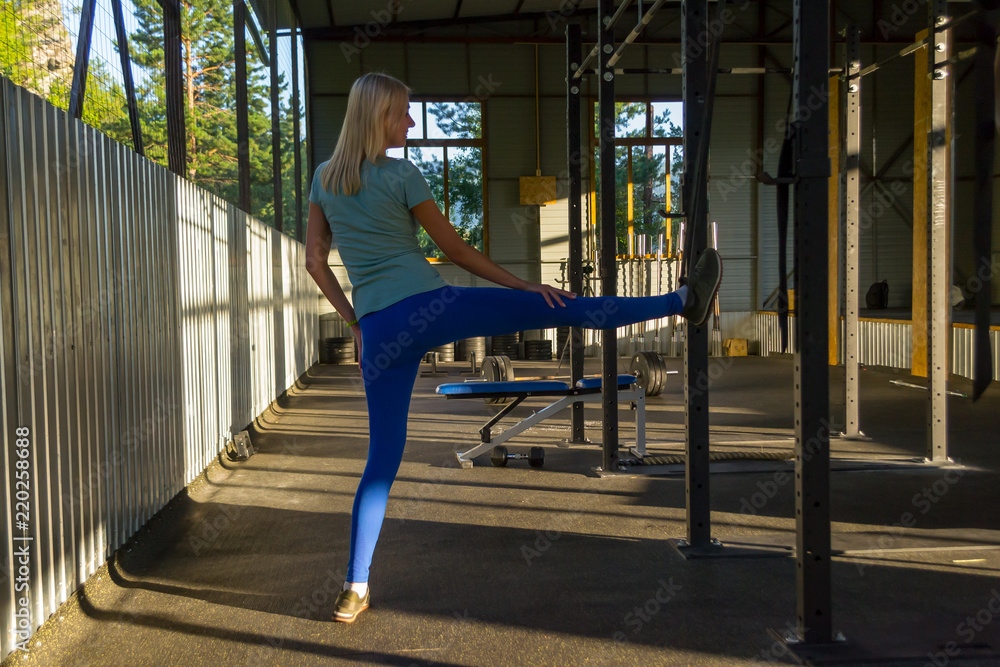 Blonde girl in blue leggings does stretching her leg up on a belt with his hands on his knee in the gym on the background of sports equipment and simulators, the windows can be seen in the mountains
