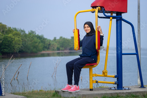Muslim woman wearing dark clothes and hijab covers her hair exercises on yellow body weight in the park near to Songkhla lake, Southern Thailand.
