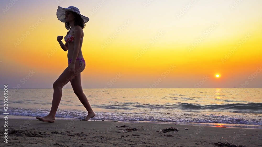 Young woman in bikini and hat walking on the beach sand in the sunset, SLOW MOTION