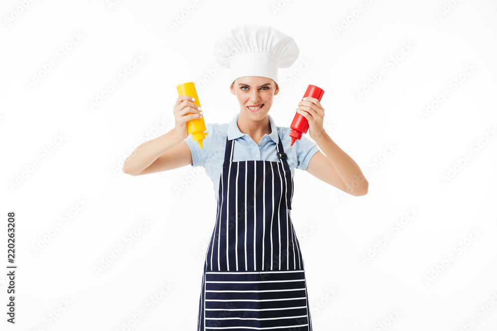 Young woman cook in striped apron and white hat playfully looking in camera while holding bottles of ketchup and mustard in hands over white background