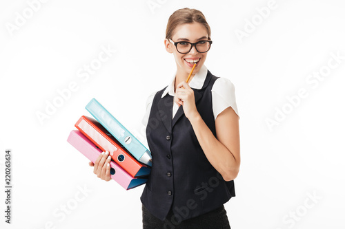 Young joyful woman in black vest and eyeglasses happily looking in camera while holding colorful folders and pencil in hands over white background