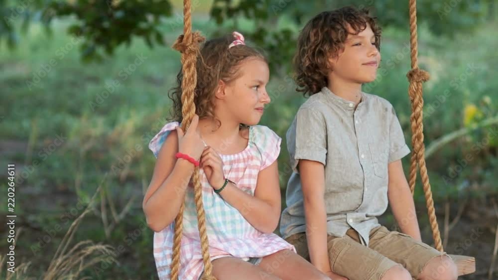 Students on vacation. A boy and a girl are rocking on a swing. Summer romantic sunset. The boy tells the girl something Students on vacation. A boy and a girl are rocking on a swing. Summer romantic sunset. The boy tells the girl something