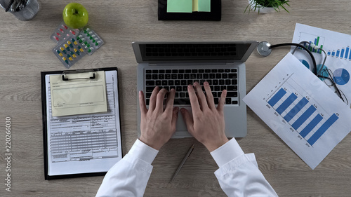 Physician looking at statistics chart, typing on laptop, preparing presentation photo