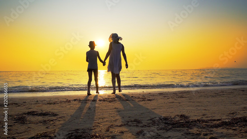 Family walk on beach to sea sunset and splash waves. Woman wear white dress and straw hat  boy has hat