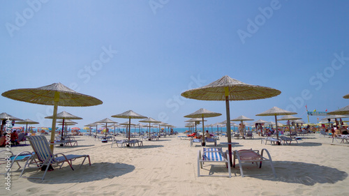 Summer beach resort bar lounge area with wooden easy chairs and straw parasols lined up next to the sea water