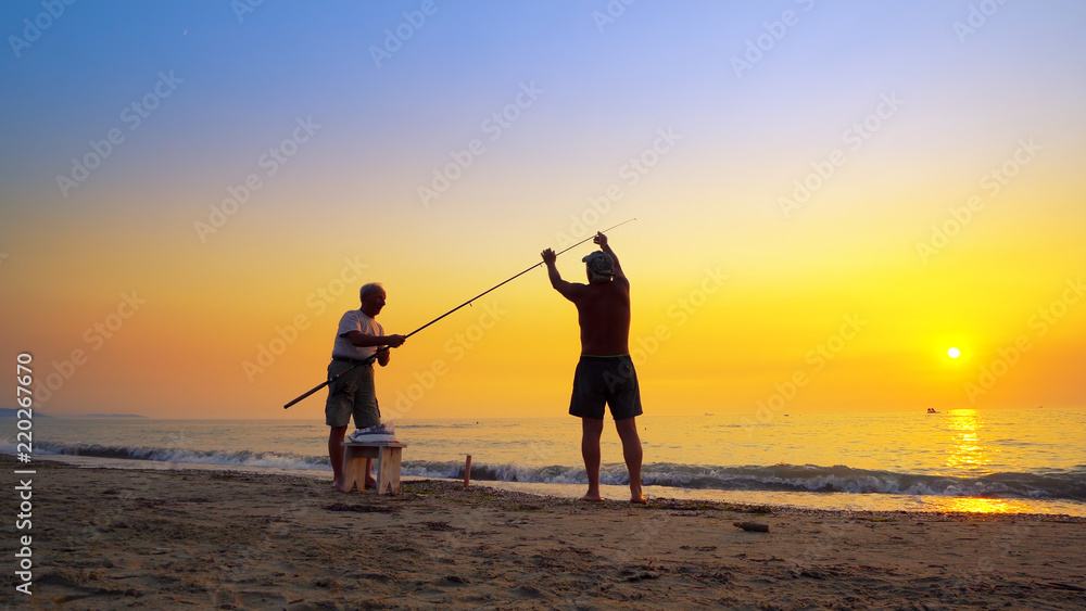 Silhouette of active fishermans fishing with rod at summer sea sunset