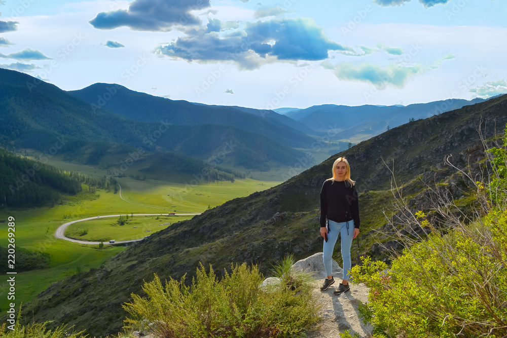 A young beautiful blonde girl stands on the edge of a cliff in the mountains of Altai on a bright sunny day, below is a green fresh grass and an asphalt road with a sharp turn leaving behind a rock