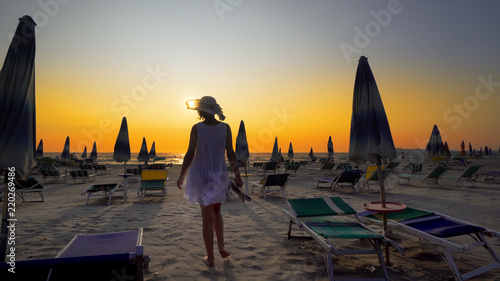 Sexy woman having fun walking, jumping, turning on empty beautiful beach with straw umbrellas and beds against sunset, cinematic shot