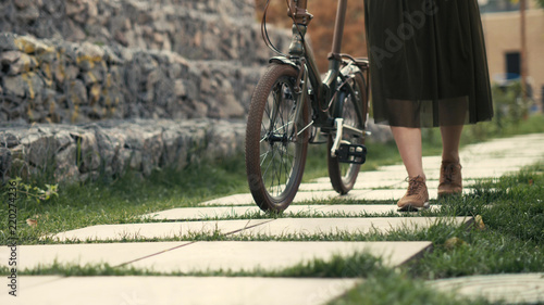 Woman legs walking beside riding bike on city road. Woman feet and bicycle wheel