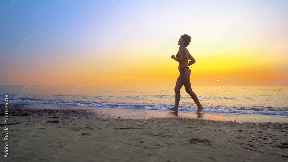 Barefoot sport woman in bikini jogging, running on an empty beach at sunset