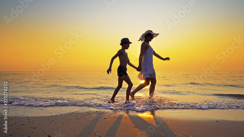 Family on summer vacation. Mother in white dress and son wearing hat walk on an epmty beach at vibrant sunset
