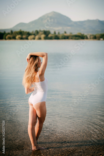 Portrait of a young stunning sexy woman in a swimsuit standing in a lake  by the sea. Beautiful slender attractive girl posing in the water at sunset. Against the mountains  