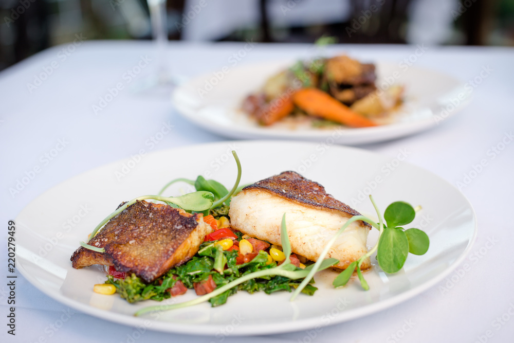 Pan roasted sable fish with spring vegetables on a white plate in a restaurant.