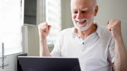Handsome elderly senior man working on laptop computer at home. Received good news excited and happy. Remote freelance work on retirement, active modern lifestyle of older people. Success concept