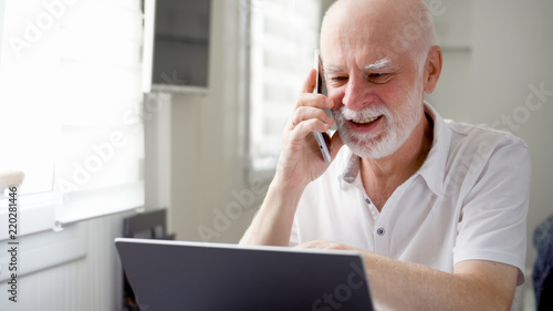 Good-looking handsome senior man sitting at home with laptop and smartphone. Using cellphone discussing project on screen. Remote freelance work on retirement, active modern lifestyle of older people