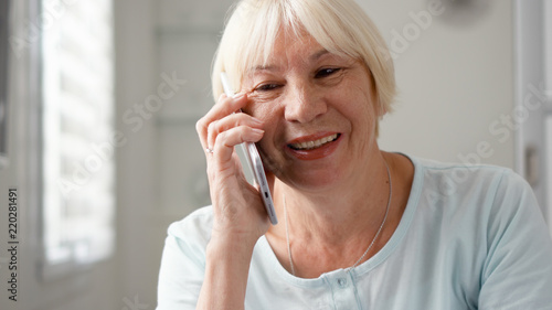 Good-looking blond senior woman sitting at home using smartphone. Retired woman talking on cellphone. Active modern elderly people concept
