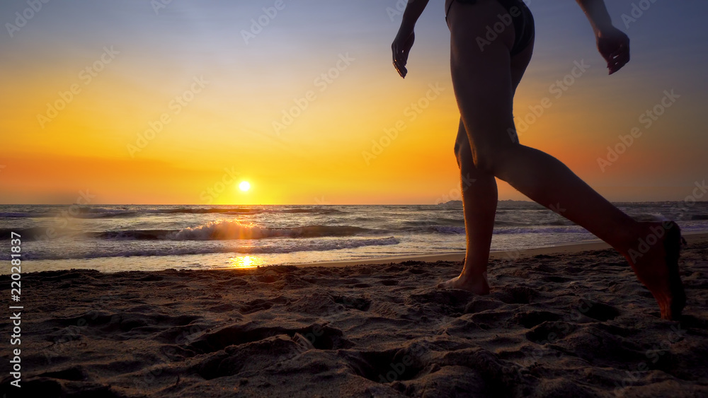 Young woman walking on a sandy beach splashed by ocean waves at sunset
