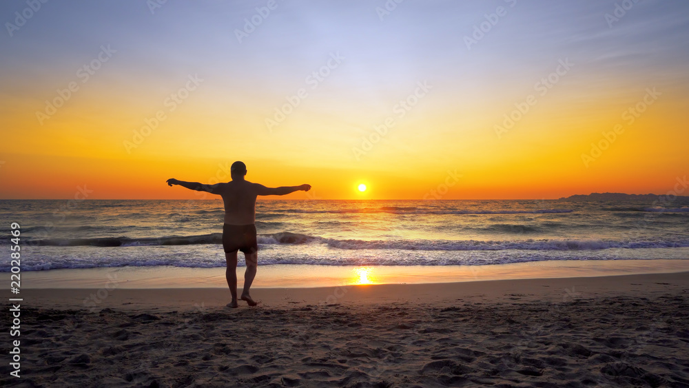 Man with hands wide open in VICTORY pose walks on empty ocean beach into water with vibrant sunset sun at background, cinematic steadicam shot
