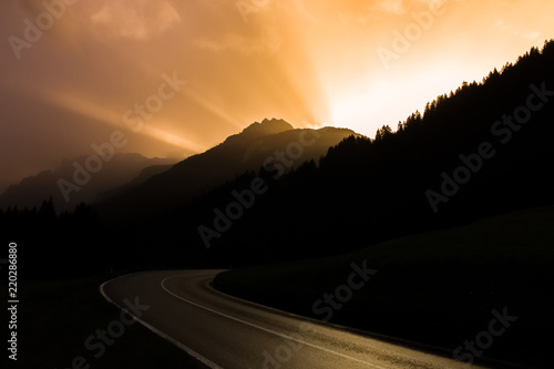 Dark road turn in a sunset with sun beams emerging from behind a mountain © Tomas Buzek