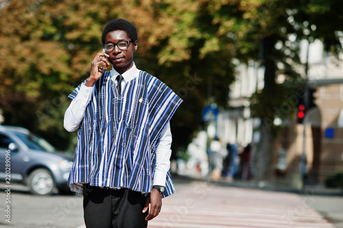 African man in traditional clothes and glasses walking at crosswalk and speaking on mobile phone.