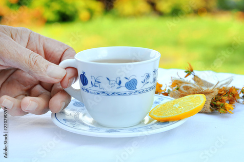 Senior woman holding cup of hot tea with lemon slice in nature.Rustic porcelain cup a herb wrapped up as a gift on a white tablecloth with lace. Healthy concept. 