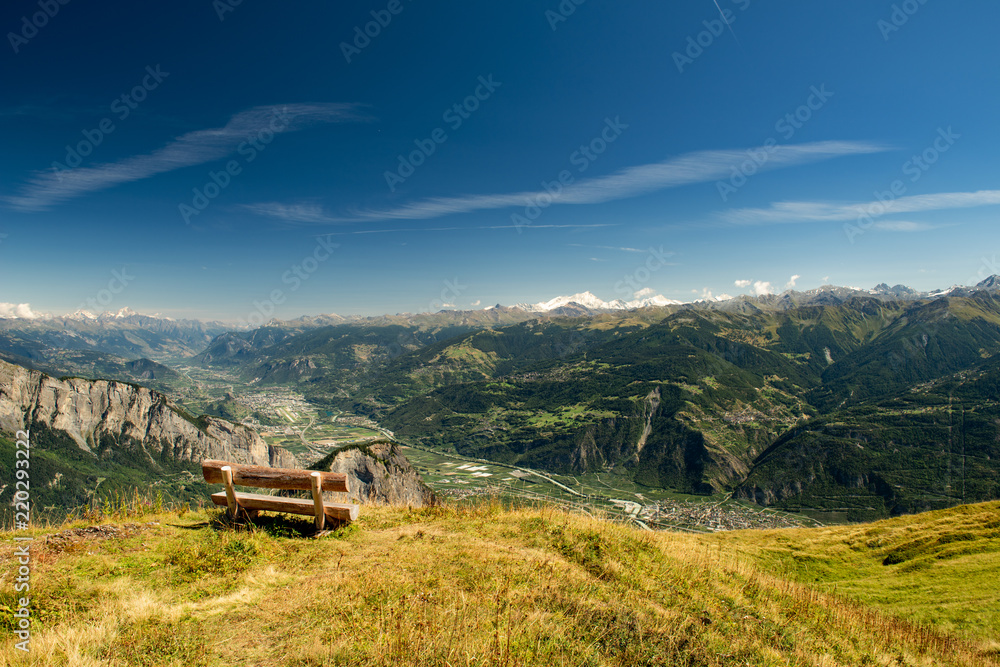 Obraz premium Scenery alpine panorama of Switzerland in sunny day with view on Ovronnaz, mountains, green forest, meadows, blue sky and white clouds with a bench for rest in the foreground