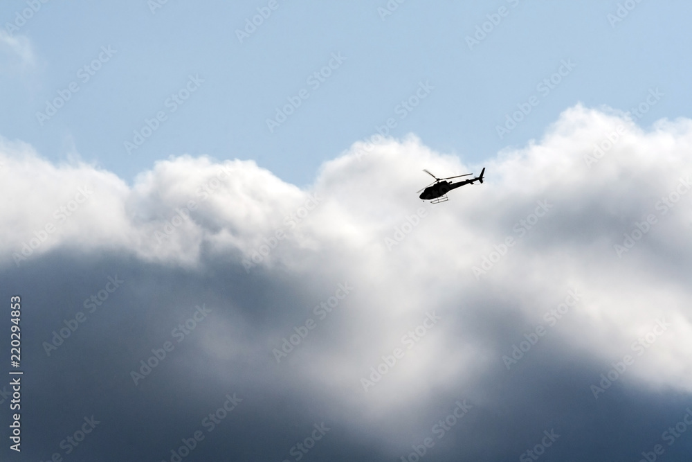 against a background of the sky of dark blue and white-blue, a black helicopter flies, clouds of white and dark clouds are combined, sunlight