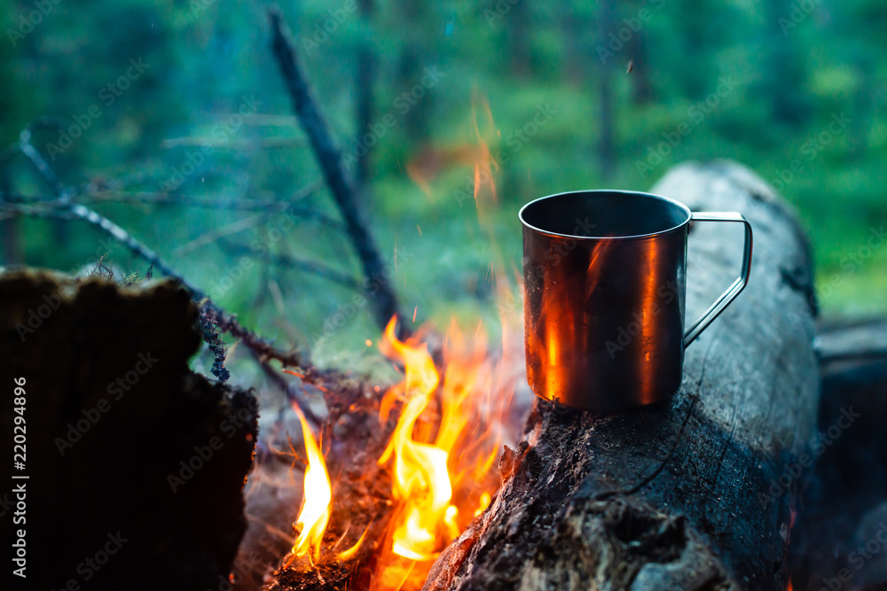 Tea in metal mug heats up on stone on bonfire. Hot drink on nature. Tea drinking in open air. Camping in dusk. Romantic warm atmosphere outdoor in twilight . Active rest. Tea in metal mug heats up on stone on bonfire. Hot drink on nature. Tea drinking in open air. Camping in dusk. Romantic warm atmosphere outdoor in twilight . Active rest.